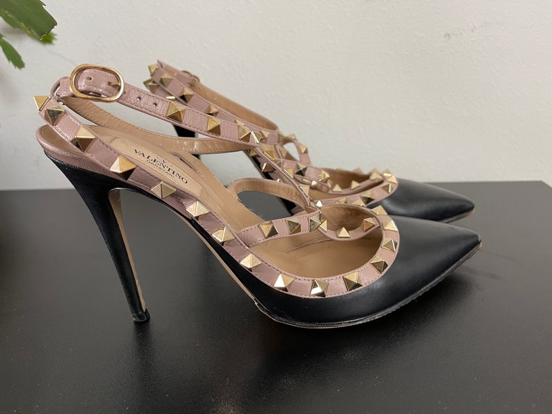 May include: Black leather high heel shoes with gold studs and a pink leather strap. The shoes have a pointed toe and a stiletto heel. The brand name "VALENTINO GARAVANI" is visible on the strap.
