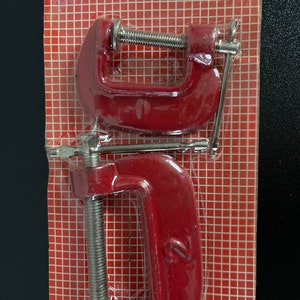 May include: Two red metal C-clamps with silver screws and handles. The clamps are packaged on a red and white grid background.