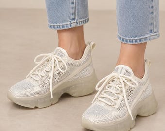 May include: A pair of white sneakers featuring a mesh upper with subtle silver glitter accents, white laces, and a thick, translucent, chunky rubber sole. The footwear has a low-top design with a pull tab at the heel and a textured knit pattern.