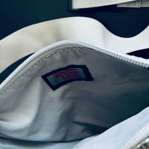 K&ouml;nnte beinhalten: Eine wei&szlig;e Rei&szlig;verschlusstasche mit einem wei&szlig;en Riemen und einem roten und wei&szlig;en Logo mit der Aufschrift "T2" im Inneren der Tasche.