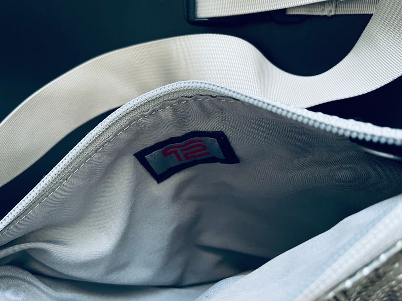 K&ouml;nnte beinhalten: Eine wei&szlig;e Rei&szlig;verschlusstasche mit einem wei&szlig;en Riemen und einem roten und wei&szlig;en Logo mit der Aufschrift "T2" im Inneren der Tasche.