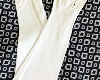 May include: A pair of long, white gloves, likely for formal wear, are displayed against a black and white geometric patterned background. The gloves are made of a smooth, likely fabric material, and have a classic design.