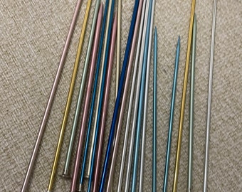 May include: A collection of 18 knitting needles in various colors, including gold, silver, blue, and pink. The needles are arranged in a fan shape on a beige surface.