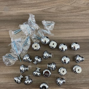 May include: A collection of 20 silver-toned cabinet knobs with a round, slightly domed shape. The knobs are arranged in a random pattern on a wood surface.