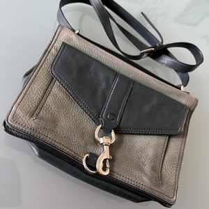 May include: A two-tone crossbody bag featuring a textured grey leather body with a smooth black leather flap. The bag includes a silver-toned metal lobster clasp closure on the front, a matching black leather shoulder strap, and black leather trim.