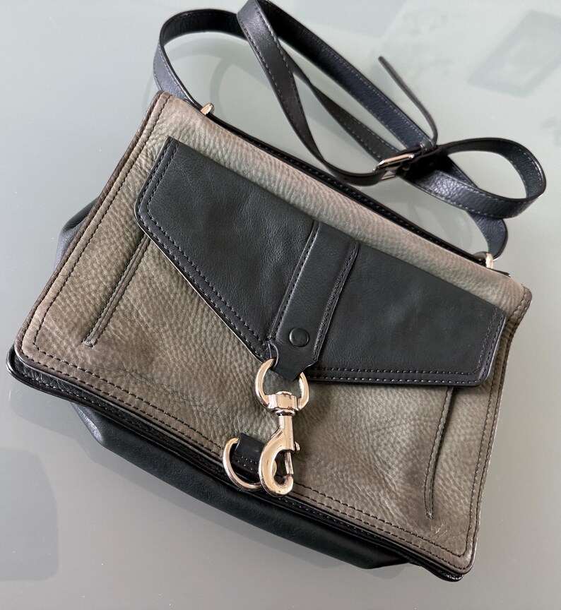 May include: A two-tone crossbody bag featuring a textured grey leather body with a smooth black leather flap. The bag includes a silver-toned metal lobster clasp closure on the front, a matching black leather shoulder strap, and black leather trim.