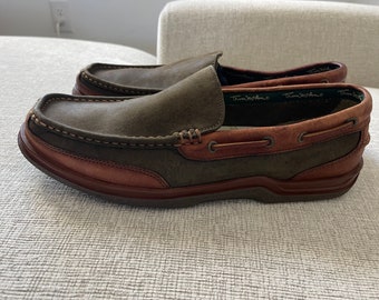 May include: Brown and green leather slip-on boat shoes with a brown leather sole and a stitched seam around the top. The shoes have a brown leather lining with the text "Toms  Womens" printed on the inside.