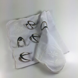 May include: Five silver napkin rings on a white tablecloth with an embroidered floral design. The napkin rings are made of a smooth, shiny metal and have a simple, elegant design.