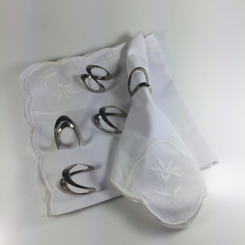May include: Five silver napkin rings on a white tablecloth with an embroidered floral design. The napkin rings are made of a smooth, shiny metal and have a simple, elegant design.