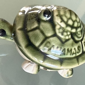 May include: A ceramic turtle figurine in shades of green, with a textured shell and visible cracks. The turtle has black eyes and the word "Bahamas" is written on the shell. The figurine is a decorative item.