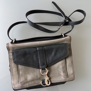 May include: A rectangular crossbody bag featuring a textured grey leather body with a smooth black leather front flap pocket. The bag includes a long, adjustable black leather shoulder strap and a prominent silver-toned metal lobster clasp closure.