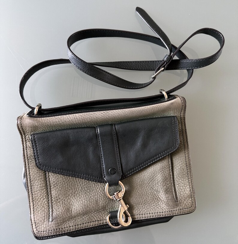 May include: A rectangular crossbody bag featuring a textured grey leather body with a smooth black leather front flap pocket. The bag includes a long, adjustable black leather shoulder strap and a prominent silver-toned metal lobster clasp closure.