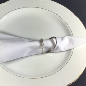 May include: A silver napkin ring with a wavy, organic design. The ring is holding a white linen napkin on a white plate with a gold rim.