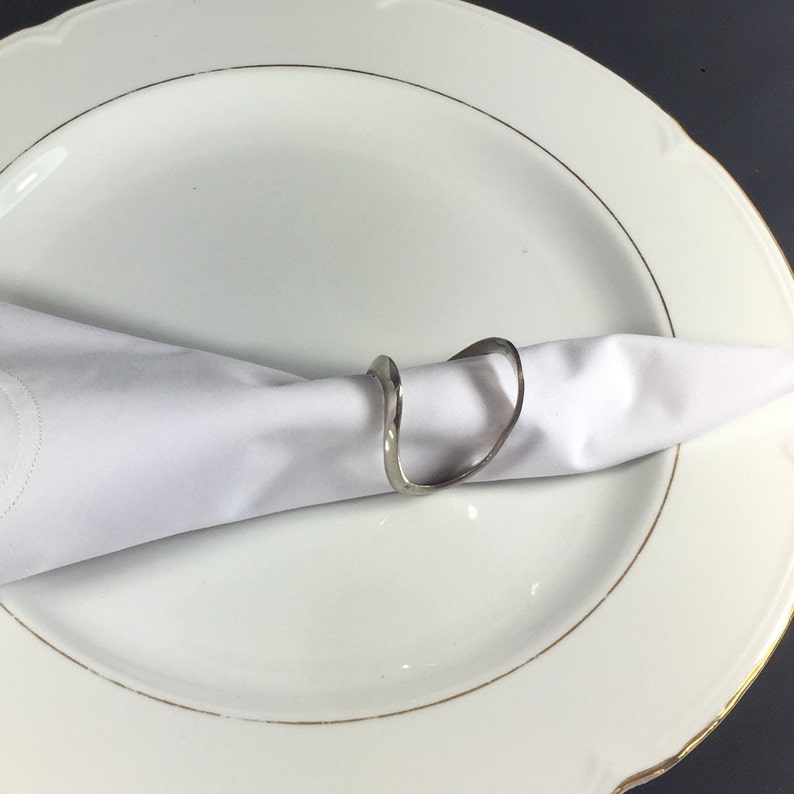 May include: A silver napkin ring with a wavy, organic design. The ring is holding a white linen napkin on a white plate with a gold rim.