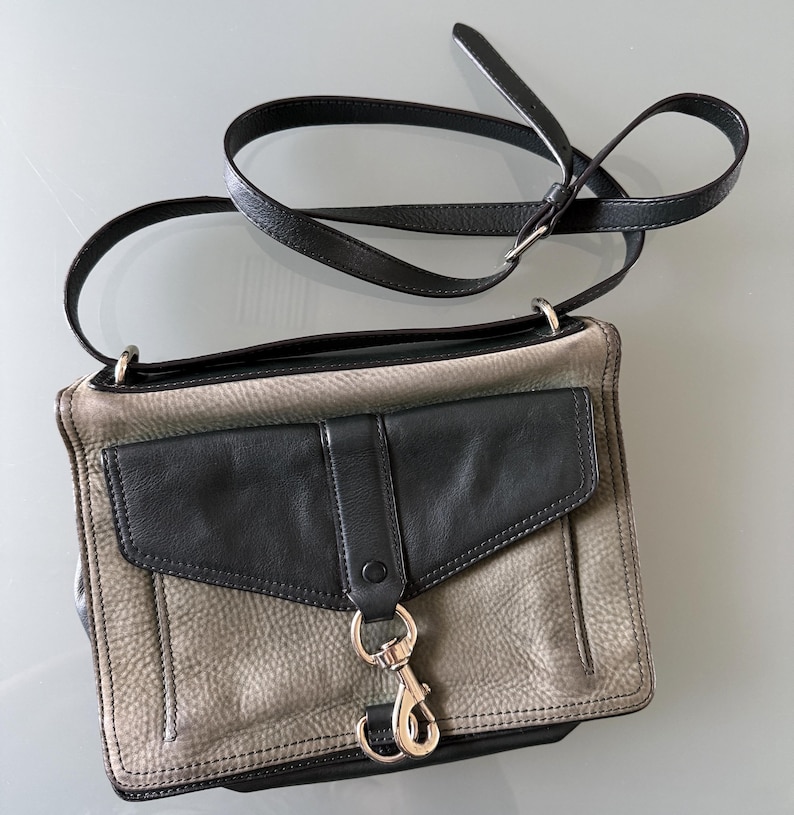 May include: A rectangular crossbody bag featuring a textured grey leather body with a smooth black leather front flap pocket. The bag includes a long, adjustable black leather shoulder strap and a prominent silver-toned metal lobster clasp closure.
