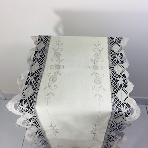 May include: A white table runner with embroidered floral designs and a delicate lace trim. The lace is black and white.