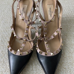 May include: Black leather heels with a pink leather strap and gold studs. The heels have a pointed toe and a low heel. The brand name "VALENTINO GARAVANI" is printed on the inside of the heel.