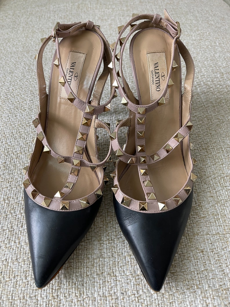 May include: Black leather heels with a pink leather strap and gold studs. The heels have a pointed toe and a low heel. The brand name "VALENTINO GARAVANI" is printed on the inside of the heel.