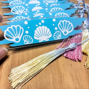 May include: Clear acrylic bookmarks with a blue seashell design and colourful tassels. The bookmarks feature white seashell graphics and bubbles on a blue background. Tassels are in shades of pink, yellow, and blue.