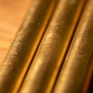 May include: Close-up of three parallel, cylindrical brass rods. The rods have a golden hue and a textured surface with visible machining marks. The background is a warm, blurred wooden surface, providing a contrast to the metallic sheen.