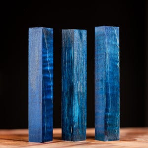 Op de afbeelding: Drie rechtopstaande, rechthoekige houten blokken in een levendige blauwe kleur. De houtnerf is zichtbaar, met lichtere strepen die een contrasterend patroon creëren. De blokken staan tegen een donkere achtergrond en benadrukken hun kleur en textuur.