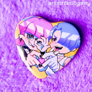 May include: Heart-shaped button featuring two anime-style characters with pink and blue hair. The characters are making a heart shape with their hands, holding a pink heart. The button has a yellow background with white outlines and the text "animatedbyamy".
