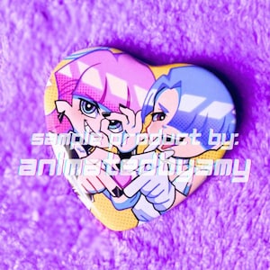 May include: A heart-shaped pin featuring a stylized illustration of two anime-style characters with pink and blue hair. The text "sample product by: animatedbyamy" is printed across the center. The background of the pin is yellow with dots.