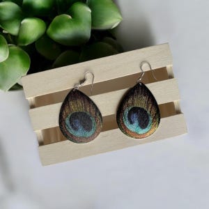 Puede incluir: Un par de pendientes en forma de lágrima con un diseño de pluma de pavo real. Los pendientes son plateados y tienen un acabado iridiscente azul, verde y dorado.