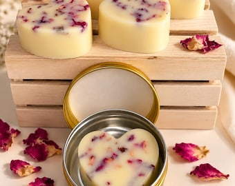Puede incluir: Cinco pastillas de jabón en forma de corazón de color blanco roto, incrustadas con pétalos de rosa secos rosas y rojos. Cuatro pastillas están dispuestas sobre una pequeña caja de madera, mientras que una se encuentra dentro de una lata redonda dorada.