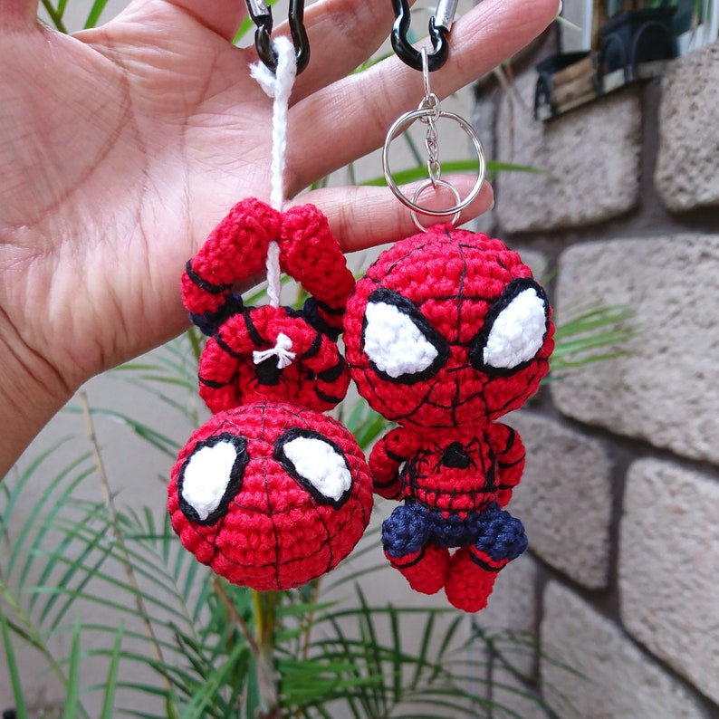 Może przedstawiać: Dwa brelokowe Spidermany wykonane na szydełku w kolorze czerwonym i czarnym. Jeden brelok to mała figurka Spidermana, drugi to głowa Spidermana.