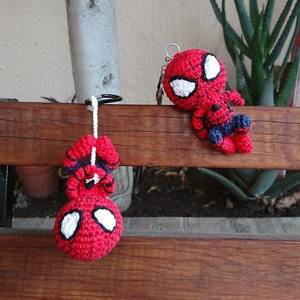 Może przedstawiać: Dwie postaci Spidermana wykonane na szydełku w kolorze czerwonym i czarnym. Jedna wisi na breloczku, a druga jest przymocowana do drewnianej powierzchni.