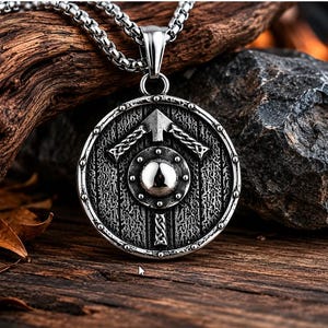 May include: A silver-colored pendant necklace featuring a round shield design with an arrow and knot details. The pendant hangs from a chain necklace. The background includes wood and stone.