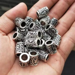 May include: A collection of silver-toned metal beads with intricate designs and runes. Each bead has a different rune symbol.