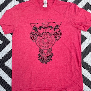 May include: Red t-shirt with a black graphic design of two skulls, a mandala, and a triangle with the text "INTER ARMA" above the design.
