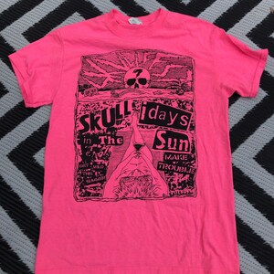 May include: Bright pink t-shirt with a black graphic design. The design features a skull, sun, and the words "Skull in the Sun" and "7 days" in a rectangular frame. The shirt is made of a soft material.