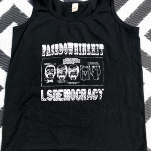 May include: Black tank top with a white graphic print that says "FACEDOWNINSHIT LSDEMOCRACY". The graphic features a series of cartoon faces and a pair of pants.