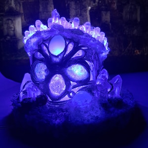 May include: A blue illuminated decorative piece, featuring a crystal crown and glowing orbs. The object is set against a blurred backdrop of a graveyard with crosses, creating a mystical and gothic aesthetic.