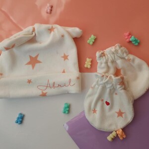May include: A white baby hat with pink stars and the name "Abril" embroidered on the front. The hat is paired with a set of white baby mittens with pink stars.