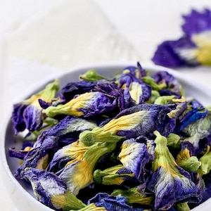 A white ceramic bowl filled with dried butterfly pea flowers. The flowers feature deep blue petals with yellow and green centers, showing a wrinkled, dehydrated texture. A single loose flower rests outside the bowl.