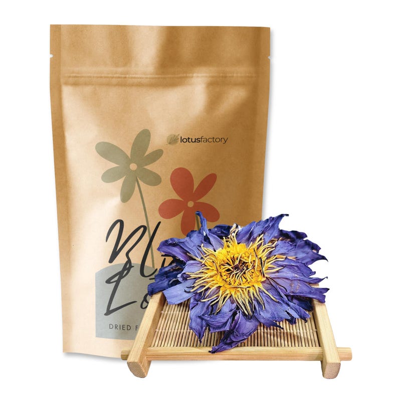 A large, dried whole blue lotus flower with vibrant purple-blue petals and a rich yellow center rests on a mini bamboo tray. Behind it stands a brown kraft paper pouch with minimalist floral branding for "lotusfactory".