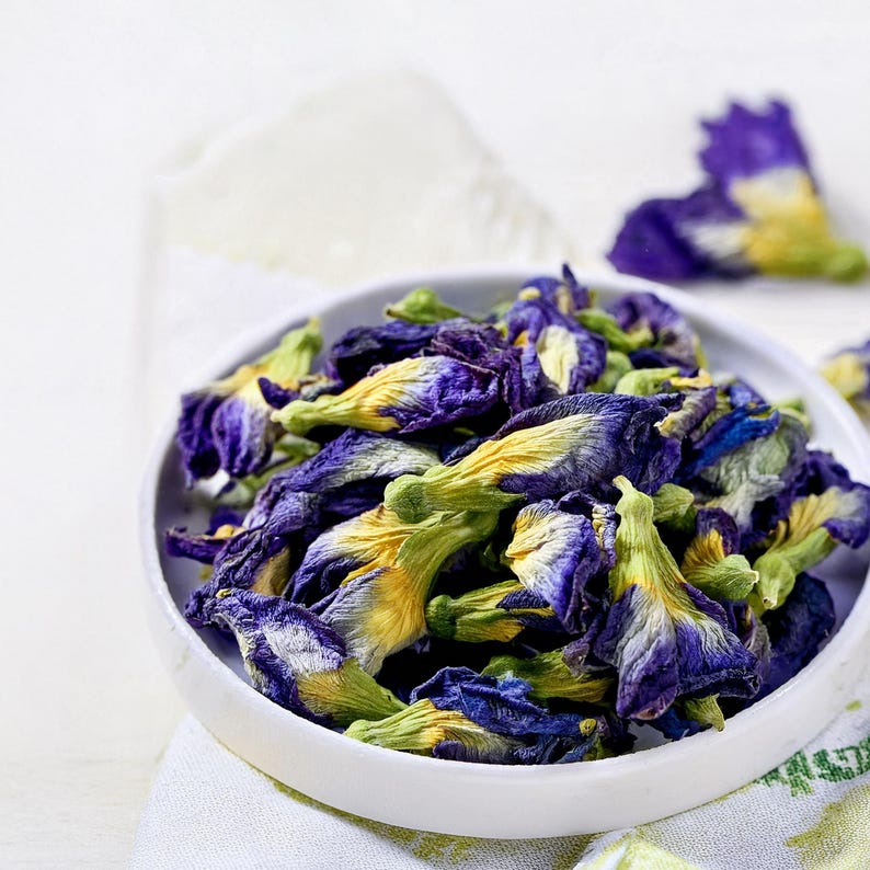 A white ceramic bowl filled with dried butterfly pea flowers. The flowers feature deep blue petals with yellow and green centers, showing a wrinkled, dehydrated texture. A single loose flower rests outside the bowl.