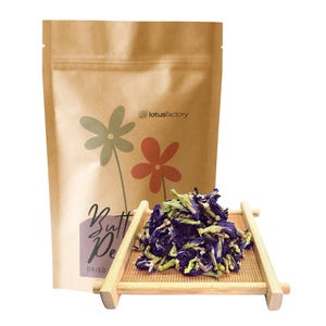 A pile of dark blue and purple dried butterfly pea flowers with green calyxes rests on a mini bamboo tray. Behind it stands a brown kraft paper pouch labeled "Butterfly Pea" with minimalist floral graphics for "lotusfactory".