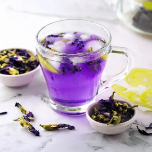 A clear glass mug filled with a vibrant purple butterfly pea flower tea, containing ice cubes and a slice of lemon. Two small bowls are filled with dried blue butterfly pea flowers, and several loose dried flowers are scattered nearby.