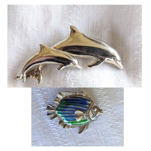 May include: Two silver-toned dolphin pins, one larger than the other, are displayed. Below, a fish pin features blue, green, and silver enamel detailing. The pins are likely jewelry or decorative accessories.