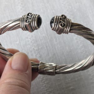May include: Silver-toned cuff bracelet with a twisted rope design. The ends feature decorative caps with black gemstones. The bracelet is held open, showcasing the clasp. A stylish piece of jewelry.