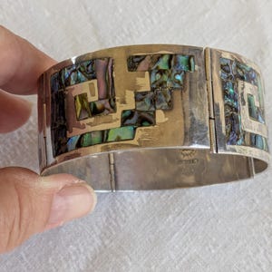 May include: A wide silver cuff bracelet featuring inlaid iridescent abalone shell segments arranged in a geometric pattern. The metal surface is polished and reflective, with visible hinges connecting the segments of the bracelet.