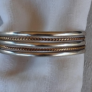 May include: A cuff bracelet featuring three smooth, polished silver-toned bands alternating with two thinner, textured, gold-toned twisted rope bands. The bracelet has a layered, multi-band design with a reflective metallic finish.