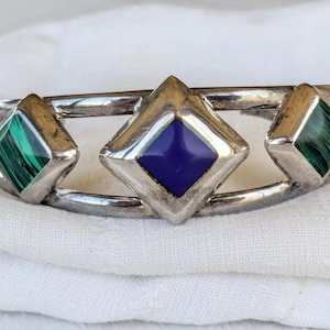 May include: Silver-toned cuff bracelet with three diamond-shaped insets. The central inset features a deep blue stone, flanked by two green malachite stones. The bracelet is displayed on a white fabric background.