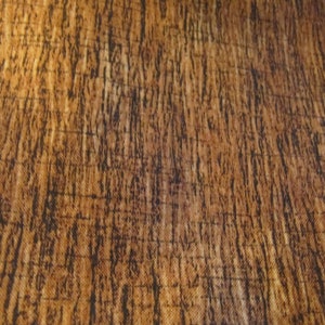 Puede incluir: Primer plano de una tela con un patrón de vetas de madera. La tela tiene una combinación de colores marrón y beige con líneas verticales marrón oscuro, creando una apariencia texturizada. El diseño imita el aspecto de la madera natural.