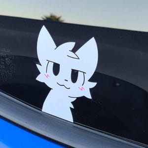 May include: A white cat sticker with a cute expression is affixed to a car window. The sticker has a simple design with a smiling face and blush marks. The car is blue.